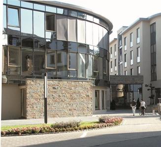 Ein modernes Gebäude mit einer geschwungenen Glasfassade und einer Steinmauer befindet sich in einer belebten Stadt, in der zwei Personen auf dem Bürgersteig gehen und eine Straßenlaterne in der Nähe steht.
